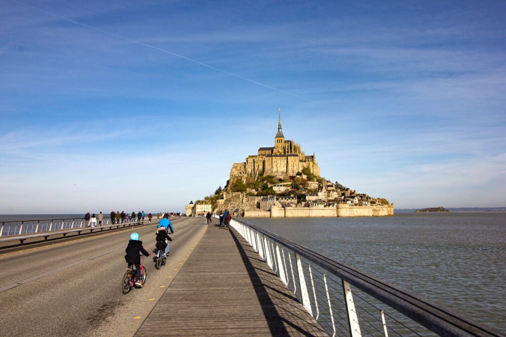 Cyclistes sur la passerelle menant au Mont, sous un ciel clair de Normandie